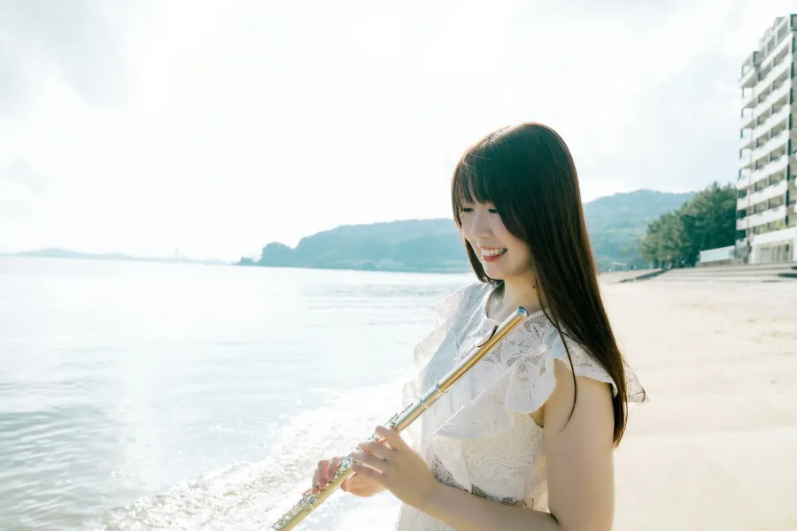 明るい曇り空と海を背景に、砂浜で笑顔でフルートを持って立つ永田梨紗の写真